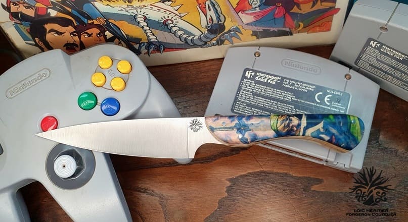 Couteau de cuisine artisanal avec un manche thématique posé sur une surface en bois. La lame droite a une finition mate et est finement affûtée pour obtenir un bord tranchant, indiquant qu'elle est conçue pour une coupe de précision. Près de la base de la lame, il y a un motif ou un logo gravé qui ajoute une touche distinctive, suggérant qu'il peut s'agir d'une signature de savoir-faire ou d'identité de marque. Le manche du couteau présente un design coloré et détaillé qui semble s'inspirer de la série de jeux vidéo Zelda, affichant des éléments pouvant être reconnus par les fans de la franchise. Le motif comprend une gamme de bleus, de verts et d'autres teintes qui forment une image complexe et vibrante, susceptible de refléter les thèmes fantastiques du jeu. En arrière-plan, au-delà de la surface en bois, se trouvent des objets qui semblent liés au jeu, notamment ce qui ressemble à une manette Nintendo 64 et à une cartouche de jeu. Ces objets soulignent le lien thématique du couteau avec le monde du jeu, en particulier avec la série Zelda, suggérant que le couteau est un objet de collection pour les passionnés qui apprécient la qualité artisanale et la culture du jeu. Le cadre général suggère un espace personnel où coexistent des souvenirs de jeu et des outils artisanaux, attirant ceux qui aiment à la fois la cuisine et les jeux.