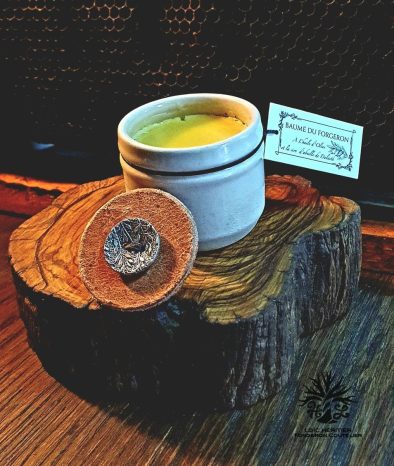 Image d'un pot de baume ouvert reposant sur une coupe transversale de tronc d'arbre, avec un couvercle en cuir embossé d'un symbole métallique, devant un fond de nid d'abeille.