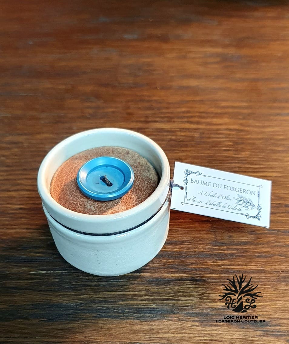 Baume du forgeron dans un pot en céramique sur une surface en bois, avec une étiquette indiquant sa composition naturelle, à côté d'un couvercle orné d'un bouton bleu.