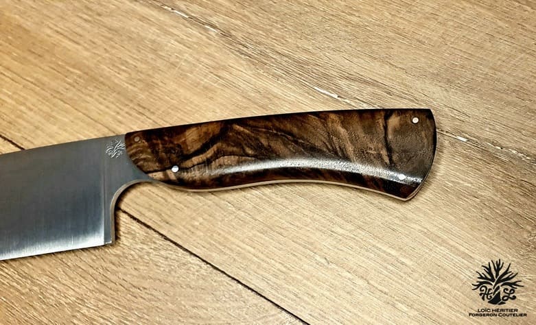 Élégance et précision définissent ce Couteau de Chef Artisanal, exposé sur un fond en bois clair qui met en valeur son esthétique raffinée. La lame, lisse et tranchante, est fabriquée en acier inoxydable X46Cr13, promettant une coupe impeccable et une résistance à la corrosion. Le manche en ronce de noyer de Provence, aux motifs tourbillonnants uniques, offre une prise en main confortable et une touche d'authenticité. La qualité de la fabrication artisanale française est mise en évidence par les rivets en acier qui accentuent l'aspect traditionnel et durable de ce couteau. Idéal pour les cuisiniers professionnels et les amateurs de gastronomie, ce couteau est un hommage à l'art culinaire.