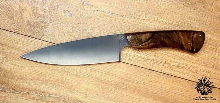 Élégance et précision définissent ce Couteau de Chef Artisanal, exposé sur un fond en bois clair qui met en valeur son esthétique raffinée. La lame, lisse et tranchante, est fabriquée en acier inoxydable X46Cr13, promettant une coupe impeccable et une résistance à la corrosion. Le manche en ronce de noyer de Provence, aux motifs tourbillonnants uniques, offre une prise en main confortable et une touche d'authenticité. La qualité de la fabrication artisanale française est mise en évidence par les rivets en acier qui accentuent l'aspect traditionnel et durable de ce couteau. Idéal pour les cuisiniers professionnels et les amateurs de gastronomie, ce couteau est un hommage à l'art culinaire.