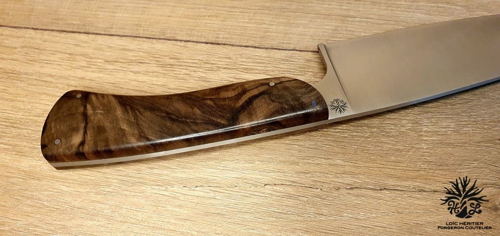 Élégance et précision définissent ce Couteau de Chef Artisanal, exposé sur un fond en bois clair qui met en valeur son esthétique raffinée. La lame, lisse et tranchante, est fabriquée en acier inoxydable X46Cr13, promettant une coupe impeccable et une résistance à la corrosion. Le manche en ronce de noyer de Provence, aux motifs tourbillonnants uniques, offre une prise en main confortable et une touche d'authenticité. La qualité de la fabrication artisanale française est mise en évidence par les rivets en acier qui accentuent l'aspect traditionnel et durable de ce couteau. Idéal pour les cuisiniers professionnels et les amateurs de gastronomie, ce couteau est un hommage à l'art culinaire.