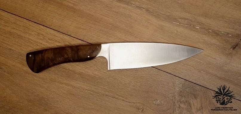 Élégance et précision définissent ce Couteau de Chef Artisanal, exposé sur un fond en bois clair qui met en valeur son esthétique raffinée. La lame, lisse et tranchante, est fabriquée en acier inoxydable X46Cr13, promettant une coupe impeccable et une résistance à la corrosion. Le manche en ronce de noyer de Provence, aux motifs tourbillonnants uniques, offre une prise en main confortable et une touche d'authenticité. La qualité de la fabrication artisanale française est mise en évidence par les rivets en acier qui accentuent l'aspect traditionnel et durable de ce couteau. Idéal pour les cuisiniers professionnels et les amateurs de gastronomie, ce couteau est un hommage à l'art culinaire.