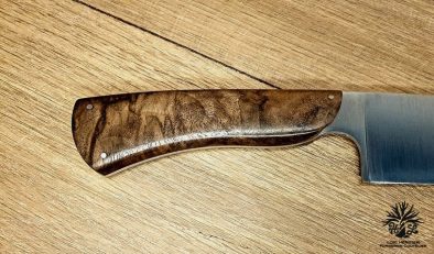 Élégance et précision définissent ce Couteau de Chef Artisanal, exposé sur un fond en bois clair qui met en valeur son esthétique raffinée. La lame, lisse et tranchante, est fabriquée en acier inoxydable X46Cr13, promettant une coupe impeccable et une résistance à la corrosion. Le manche en ronce de noyer de Provence, aux motifs tourbillonnants uniques, offre une prise en main confortable et une touche d'authenticité. La qualité de la fabrication artisanale française est mise en évidence par les rivets en acier qui accentuent l'aspect traditionnel et durable de ce couteau. Idéal pour les cuisiniers professionnels et les amateurs de gastronomie, ce couteau est un hommage à l'art culinaire.
